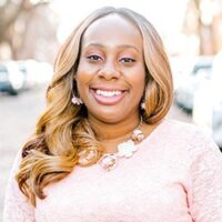 A smiling woman in a pink dress standing on a street named Danielle Blackshear.