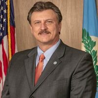 A man in a suit and tie standing in front of flags at a Tim Gatz event.